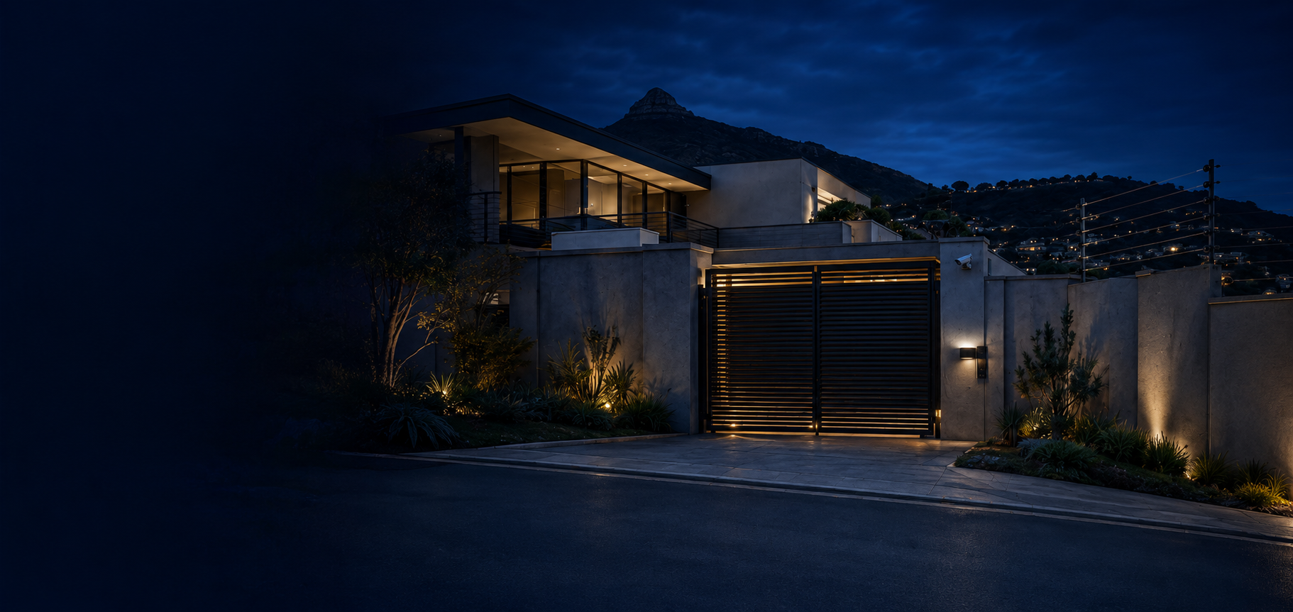 Modern hillside home at night with an automated gate, security camera and electric fencing along the boundary wall